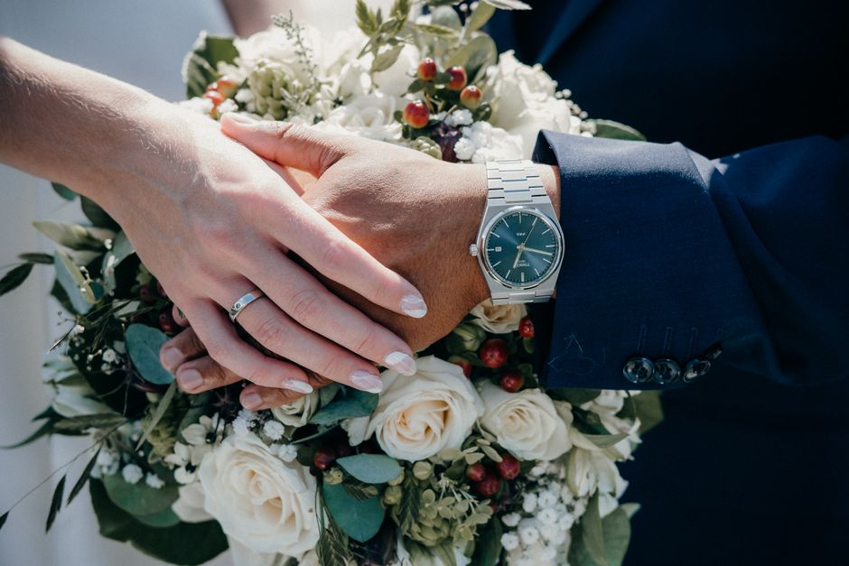 A close-up image of a newlywed couple's hands, with the bride's hand resting atop the groom's, both displaying wedding rings. The bride's hand has a simple silver band, and her nails are neatly manicu