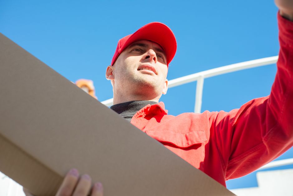 A delivery person wearing a red uniform and red cap is holding a cardboard box, possibly containing floral arrangements, with a clear blue sky in the background. The individual is looking slightly upw