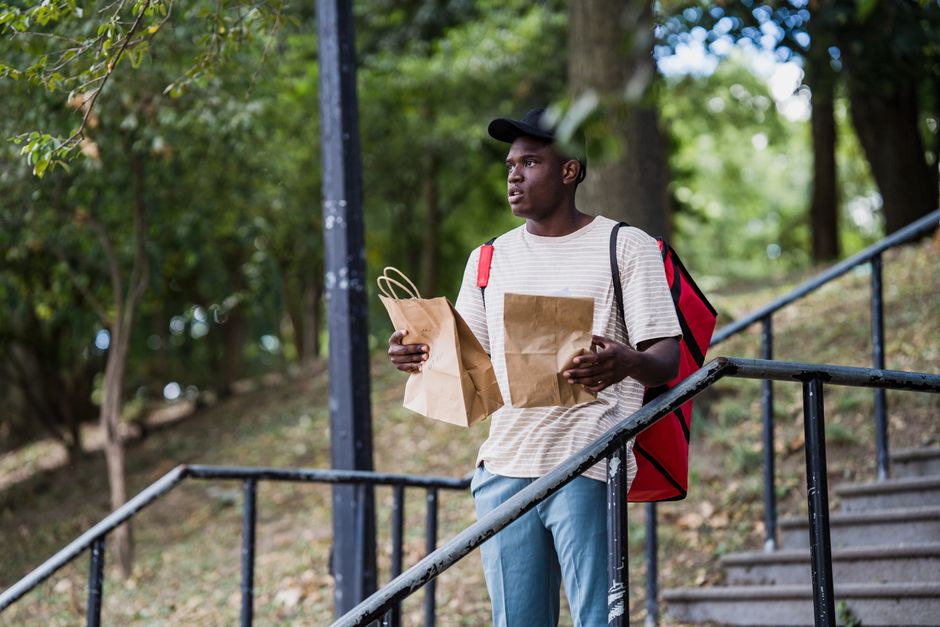 A young man with dark skin and a serious expression stands outdoors on a metal staircase, surrounded by lush green trees and foliage. He is holding two brown paper gift bags, one in each hand, and is
