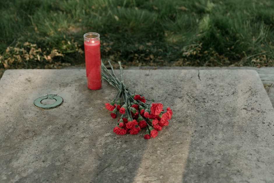 A small memorial tribute consisting of a bunch of red carnations with green stems, laid gently on a weathered stone surface. Next to the flowers is a partially filled red memorial candle in a glass co