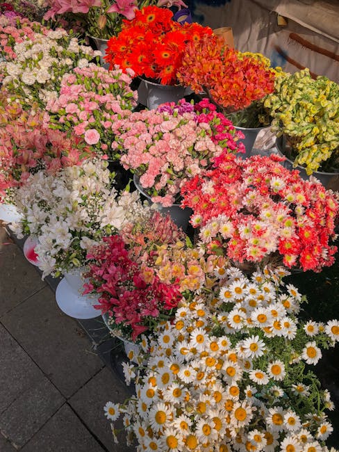 A vibrant display of freshly cut flowers arranged in buckets and bowls, featuring a variety of blooms including pink and white chrysanthemums, white daisies with yellow centers, bright red and orange 