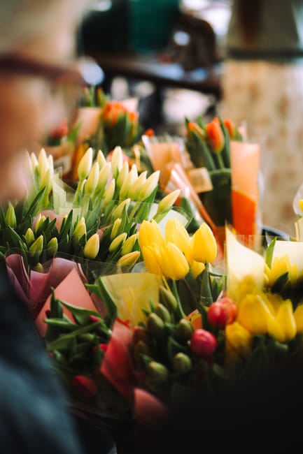 A vibrant display of freshly cut tulips and other spring flowers at a floral shop, featuring bright yellow, white, and pink blooms arranged in clear wrapping paper and decorative tissue paper. The flo